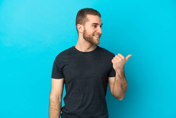 Jonge knappe blanke man geïsoleerd op blauwe achtergrond wijzend naar de zijkant om een product te presenteren - Foto, afbeelding