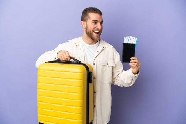 Молодой красивый белый мужчина изолирован на фиолетовом фоне в отпуске с чемоданом и паспортом - Фото, изображение