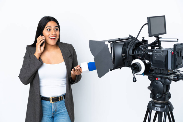 Журналістка колумбійська жінка тримає мікрофон і повідомляє новини на білому тлі про те, що веде розмову з кимось на мобільному телефоні. - Фото, зображення
