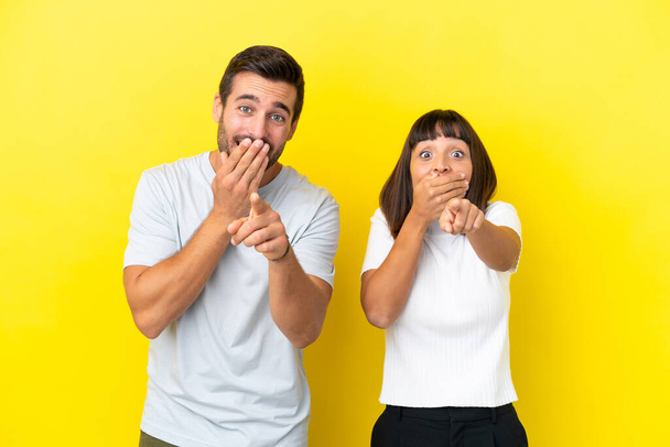Молодая пара изолирована на желтом фоне, указывая пальцем на кого-то и смеясь - Фото, изображение