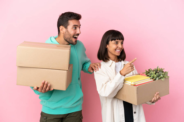 ピンクの背景に孤立した箱の中で新しい家に引っ越して製品を提示するために側面を指している若い美しさのカップル - 写真・画像
