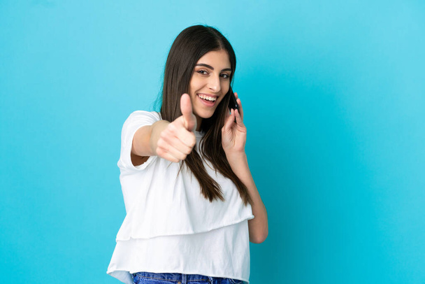 Young caucasian woman isolated on blue background keeping a conversation with the mobile while doing thumbs up - Photo, Image