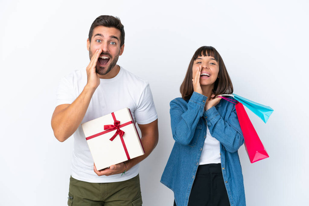 Junges Paar mit Einkaufstaschen und Geschenk isoliert auf weißem Hintergrund schreit und kündigt etwas an - Foto, Bild