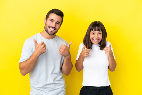 Young couple isolated on yellow background giving a thumbs up gesture with both hands and smiling - Φωτογραφία, εικόνα