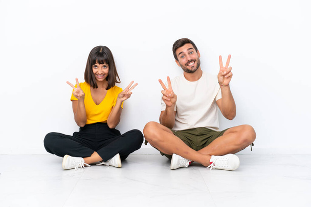 Mladý pár sedí na podlaze izolované na bílém pozadí s úsměvem a ukazuje vítězství znamení s oběma rukama - Fotografie, Obrázek