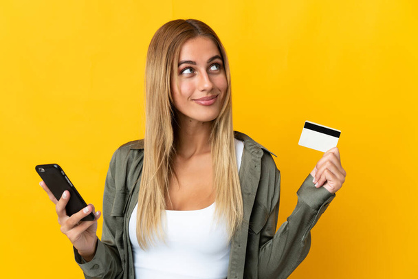 Junge blonde Frau isoliert auf gelbem Hintergrund kauft mit dem Handy mit einer Kreditkarte, während sie denkt - Foto, Bild