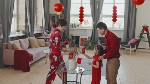 Μέτριο πλάνο της ευτυχισμένης ασιατικής οικογένειας με 5-year-old κόρη φορώντας παραδοσιακά ρούχα και βάζοντας κινέζικα στολίδια Πρωτοχρονιάς σε κλαδιά στο βάζο στο σαλόνι διακοσμημένα με χάρτινα φανάρια - Πλάνα, βίντεο
