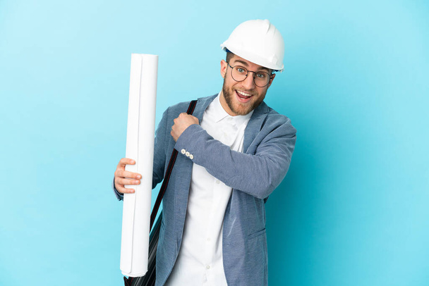 Young architect man with helmet and holding blueprints over isolated background celebrating a victory - Foto, Bild