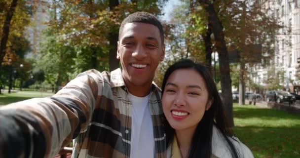 Cheerful handsome African guy holding a mobile phone in outstretched hand, tenderly hugging his pretty girlfriend and having a video conference while walking together on the urban park at sunny day - Footage, Video