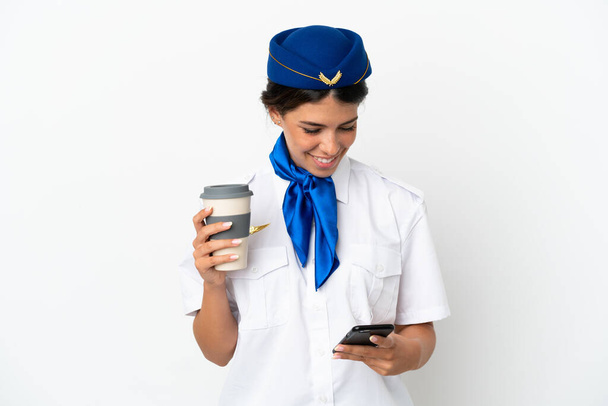 Aeromoça caucasiana mulher isolada no fundo branco segurando café para levar e um celular - Foto, Imagem