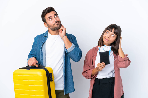 Young traveler couple holding a suitcase and passport isolated on white background thinking an idea while scratching head - Foto, Bild