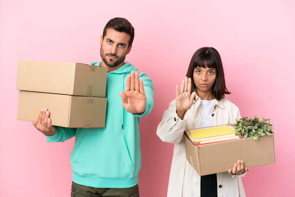 joven pareja de belleza moviéndose en nuevo hogar entre cajas aisladas sobre fondo rosa haciendo stop gesture negando una situación que piensa mal - Foto, Imagen