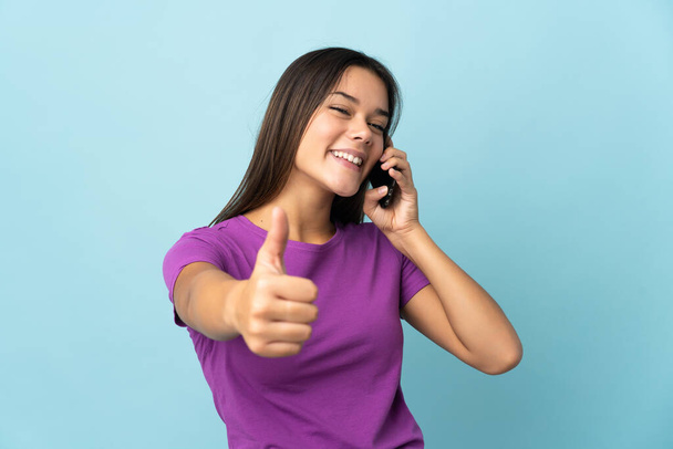 Teenager girl isolated on pink background keeping a conversation with the mobile while doing thumbs up - Fotó, kép