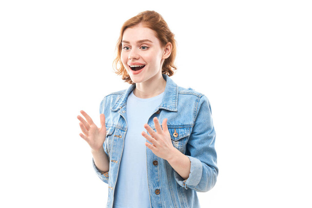 Surprise young red-haired woman enjoys looking at the camera. Smiling girl in jeans on white background looking away. Success. Spreads his arms to the sides. - Photo, Image
