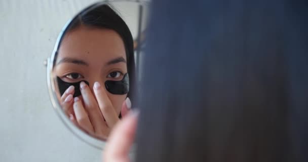 定期的なスキンケア。バスルームに立つ陽気若いアジアの美しい女性,目の下に黒の平滑化と保湿コラーゲンパッチを適用する小さな化粧鏡で彼女の反射を見て - 映像、動画