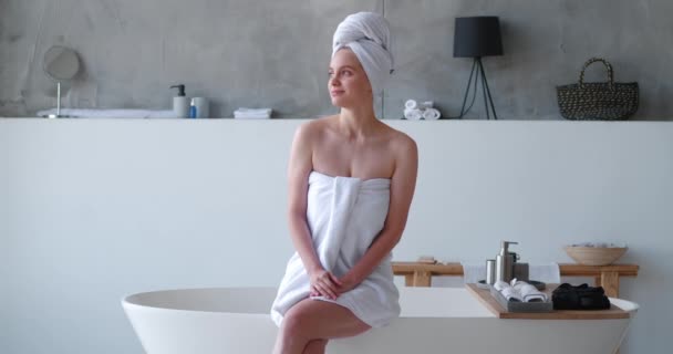 Schöne Frau in weißes Badetuch gehüllt sitzt mit überkreuzten nackten Beinen am Rand der modernen Badewanne im Luxus-Badezimmer, lächelt zuversichtlich in die Kamera, bevor sie entspannendes Schaumbad nimmt - Filmmaterial, Video