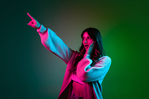 Studio shot of young adorable cute girl, student in casual style clothes posing isolated on dark green studio backgroud in pink neon light. - 写真・画像