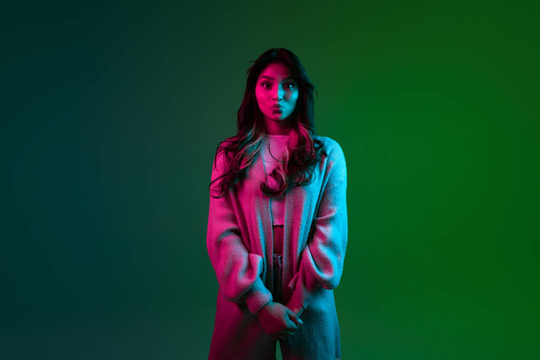 Studio shot of young adorable cute girl, student in casual style clothes posing isolated on dark green studio backgroud in pink neon light. - Zdjęcie, obraz