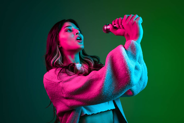 Studio shot of young adorable cute girl, student in casual style clothes posing isolated on dark green studio backgroud in pink neon light. - Foto, imagen