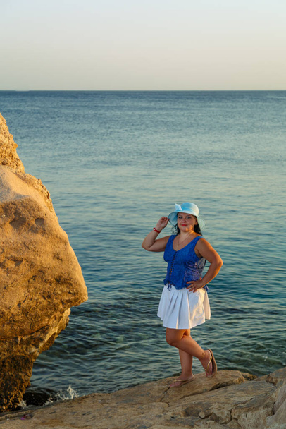 A young brunette woman in a white skirt and a sun hat on the seashore stands on a rock. - Foto, Bild