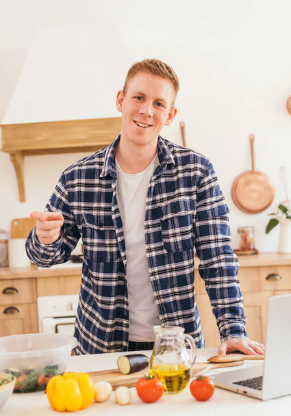 Cheerful young fair-haired guy in a plaid shirt cooks using with a laptop peeping a recipe on the internet. - Photo, Image