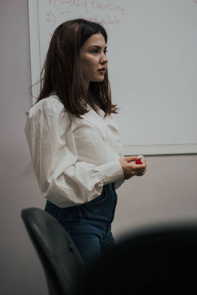 Young female english language teacher with a red marker in a white blouse standing in front of a white board - Photo, Image