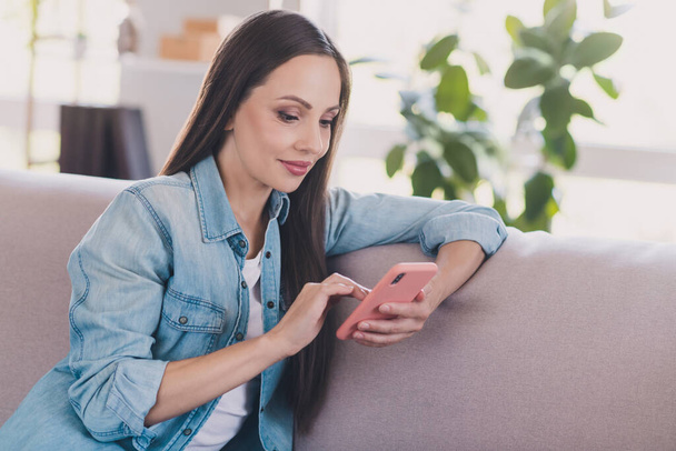 Retrato de mujer atractiva enfocada alegre influencer sentado en diván usando dispositivo post smm en casa en interiores - Foto, imagen