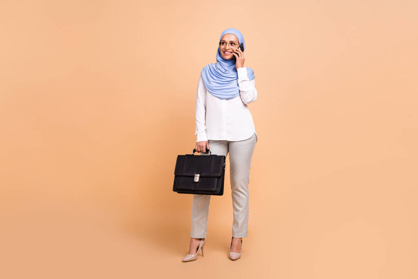 Full body photo of millennial arabic modern lady with bag talk telephone wear headscarf shirt eyewear pants stilettos isolated on beige background - Zdjęcie, obraz