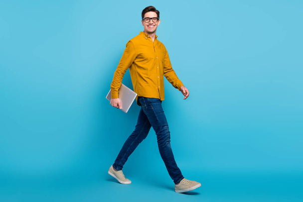 Full size profile photo of funny brunet boss man hold laptop go wear yellow shirt eyewear jeans shoes isolated on blue color background - Fotoğraf, Görsel