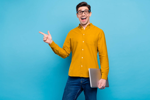 Photo of excited guy hold laptop indicate forefinger empty space wear yellow shirt isolated blue color background - Fotó, kép