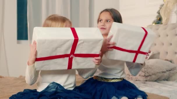İki küçük kız ellerinde Noel hediyeleri kutularıyla. Kutuda ne olduğunu öğrenmek umuduyla hediye kutularını sallayan çocukların mutlu duyguları. - Video, Çekim