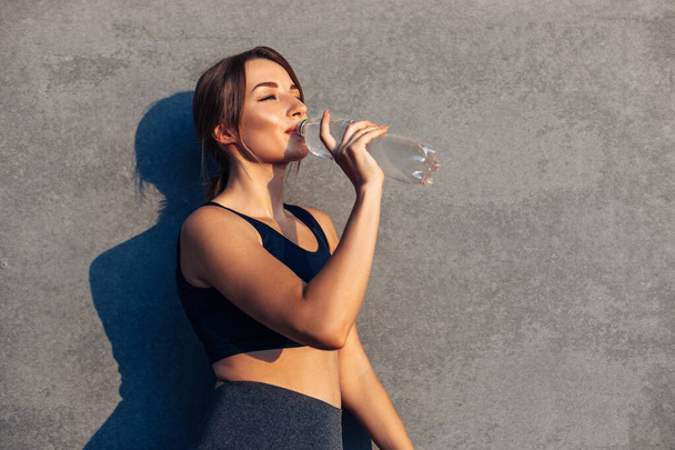 Sports fitness woman , resting after grueling jogging workout, sportswoman drinks water outdoors after workout, on isolated gray background of city street wall - Foto, Bild