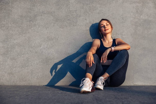 Beautiful smiling fitness woman, in sportswear, happy woman sitting on the floor against the background of a gray city wall, concept, sport, motivation - 写真・画像
