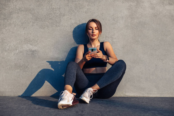 Sports fitness woman using her mobile phone during a break from workout while sitting on a gray background in the street, sport and healthy lifestyle - Fotografie, Obrázek
