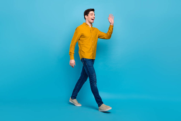 正の素敵な男のプロフィール写真歩く街波手は黄色のシャツジーンズスニーカー隔離された青の色の背景を着用 - 写真・画像