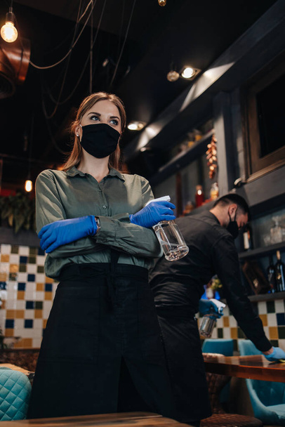 Young restaurant workers waiters cleaning and disinfecting tables and surfaces against Coronavirus pandemic disease. They are wearing protective face masks and gloves. - Foto, Imagen