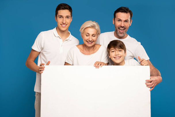 A wonderful happy family wearing white tshirts- two sons, mom and dad. Smiling, friendly people pose in the studio, holding a white empty board in their hands. Blue background. Real people family lifestyle - Photo, Image