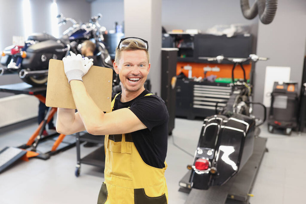 Homem feliz reparador segurando prancheta com documentos perto de motocicleta na oficina de reparação de automóveis - Foto, Imagem