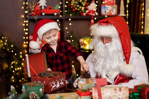Santa Claus with little boy assistant packing Christmas gifts for children. NEW YEAR CONCEPT - Фото, зображення