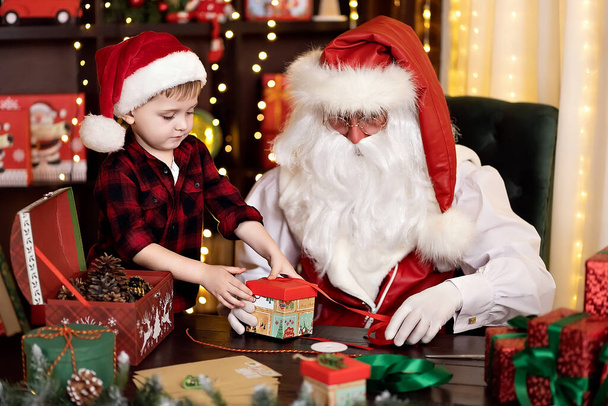 Santa Claus with little boy assistant packing Christmas gifts for children. NEW YEAR CONCEPT - Fotó, kép
