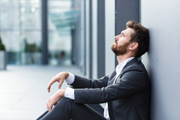 近代的なオフィスビジネスセンターの近くの屋外通りに座って正式なスーツ労働者の男で悲しい落ち込んで起業家。動揺の男性実業家は財政危機の従業員のために仕事を失った問題がある。外で - 写真・画像