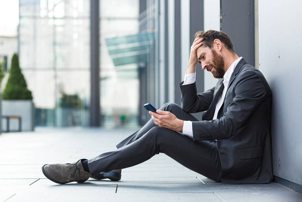 Office worker sitting near office outdoors got bad news, depressed and tired reads bad news from phone online - Foto, Bild