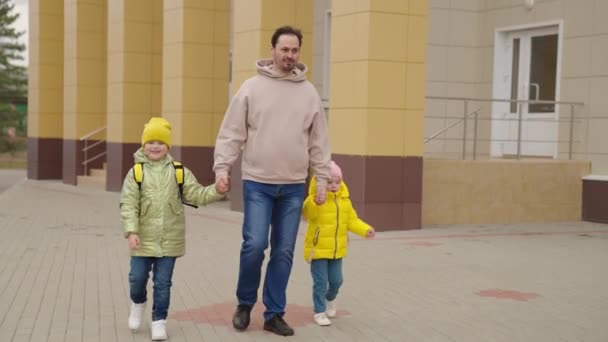 father leads children with backpacks by hand through schoolyard, educate first grade students, little kids rush lesson, baby preschool education, pick up their daughters from preschool education - Footage, Video