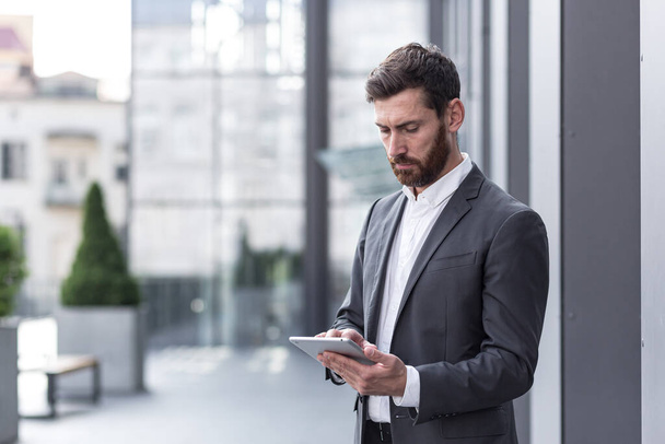 stylish bearded businessman in formal business suit standing working with tablet in hands on background modern office building outside. - Foto, immagini