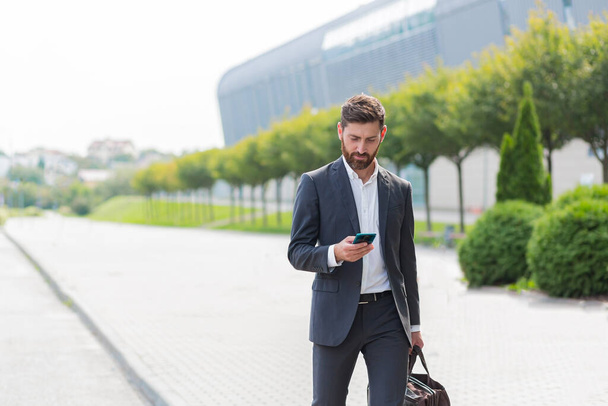 stylish bearded business man with travel bag and mobile phone in hands walks on city street modern urban background. Businessman in a suit goes and uses a smartphone. tourist trip. caucasian manager - Фото, зображення