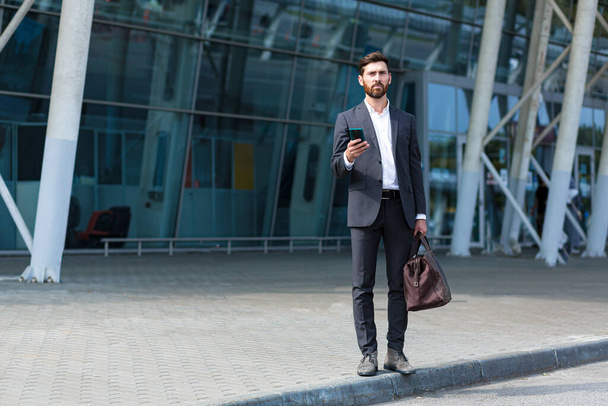 stylish bearded business man with travel bag and mobile phone in hands walks on city street modern urban background. Businessman in a suit goes and uses a smartphone. tourist trip. caucasian manager - Φωτογραφία, εικόνα