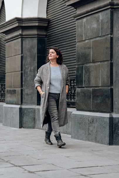 Vertikales Bild einer modischen Frau, die mit den Händen in den Hosentaschen eine Straße hinuntergeht und den Spaziergang lächelnd genießt - Foto, Bild
