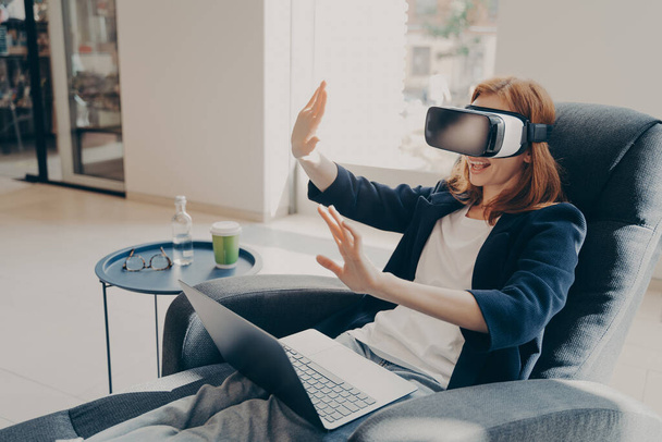Young businesswoman wearing VR headset glasses and laptop sitting in cozy armchair in office lobby - Foto, Imagem