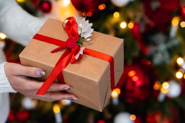 Kobiece ręce z pudełkiem prezentów w pobliżu choinki, zbliżenie. Prezent w rękach w dekoracji noworocznej - Zdjęcie, obraz