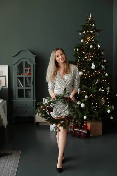 Blonde model girl in a short fashionable dress keeps in her hands a Christmas wreath next to a decorated Christmas tree - 写真・画像